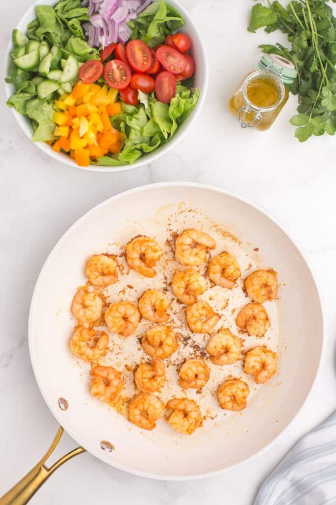 Skillet with shrimp cooking in it. Prepped salad in a large serving bowl next to the skillet and a jar of homemade dressing.
