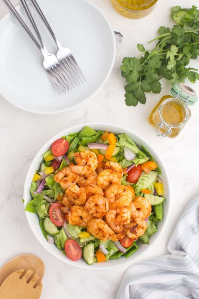 Shrimp salad in a serving bowl surrounded by a glass jar of homemade dressing, a white dinner plate with forks on it, and a set of serving tongs.