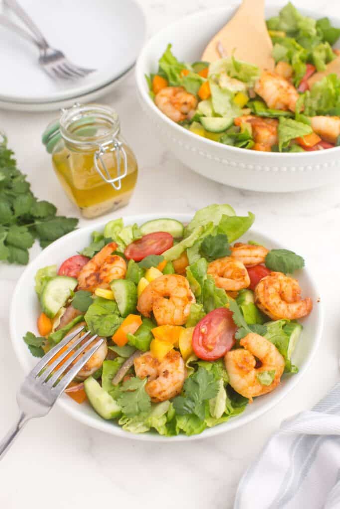 White dinner bowl with a serving of shrimp salad. A larger serving bowl is behind it with salad. A small glass container holds a homemade dressing.