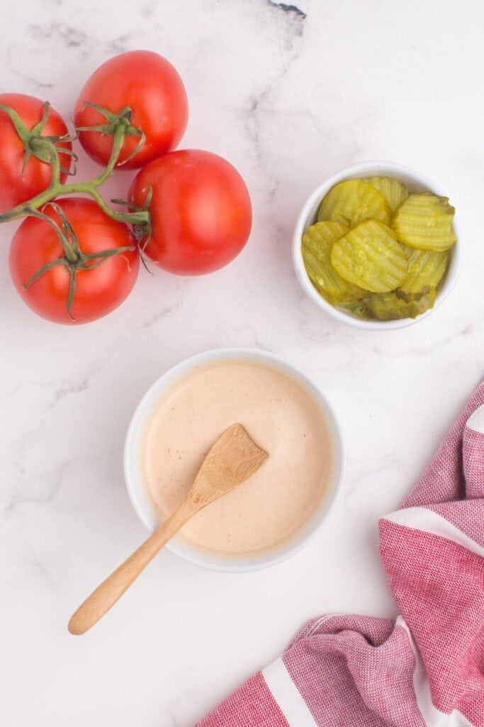 Hamburger sauce in a small white bowl with a wooden spoon in it. Bowl is surrounded by tomatoes on the vine and a bowl of pickles.