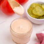 Hamburger sauce in a glass jar, surrounded by tomatoes on the vine and a bowl of pickles. A wooden spoon is lifting some sauce from the glass jar.