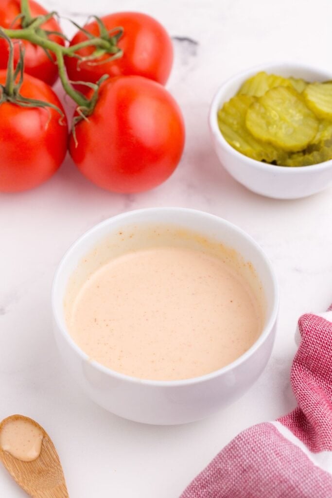Hamburger sauce in a small bowl, surrounded by tomatoes on the vine, a bowl of pickles, and a wooden spoon.