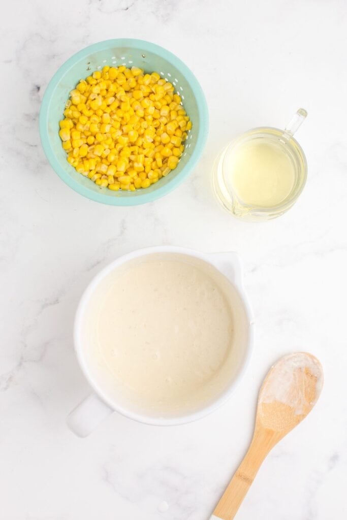 White mixing bowl with a batter in it. Next to the bowl is a colander of canned corn nibs and a pitcher of oil.