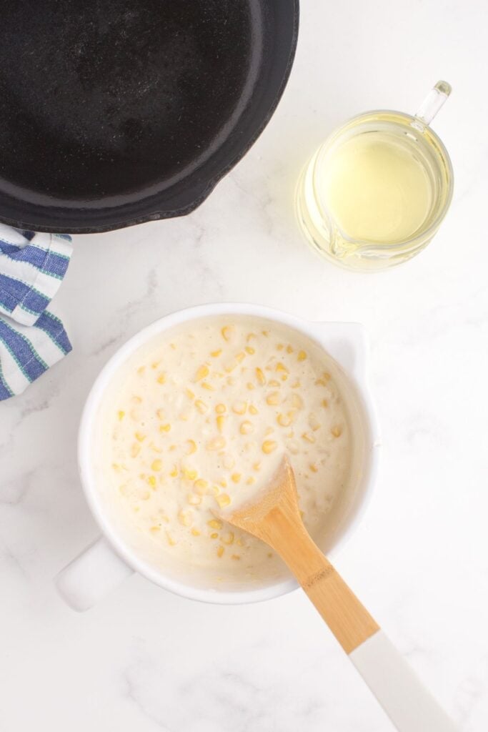 A white mixing bowl with prepared corn nugget batter in it. Mixing bowl is next to a cast iron pan and glass pitcher with oil in it.