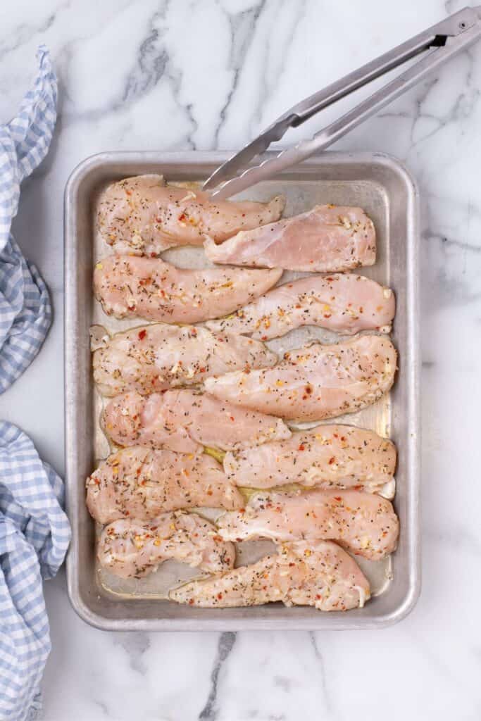 Raw chicken tenders prepped and seasoned on a quarter sheet pan.