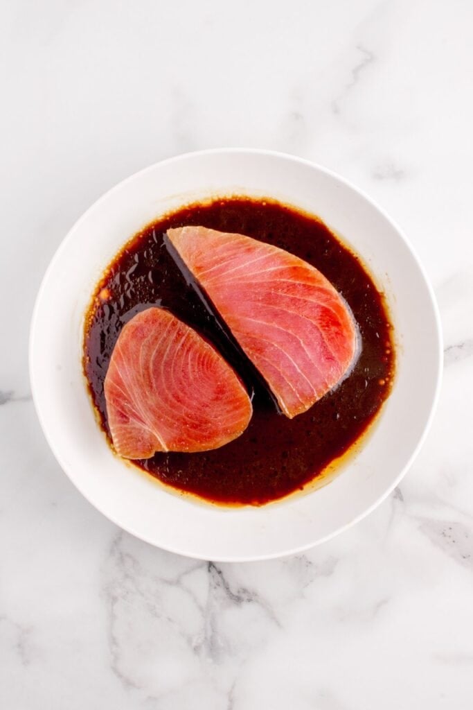 Tuna steaks resting in a marinade on a white dinner plate.
