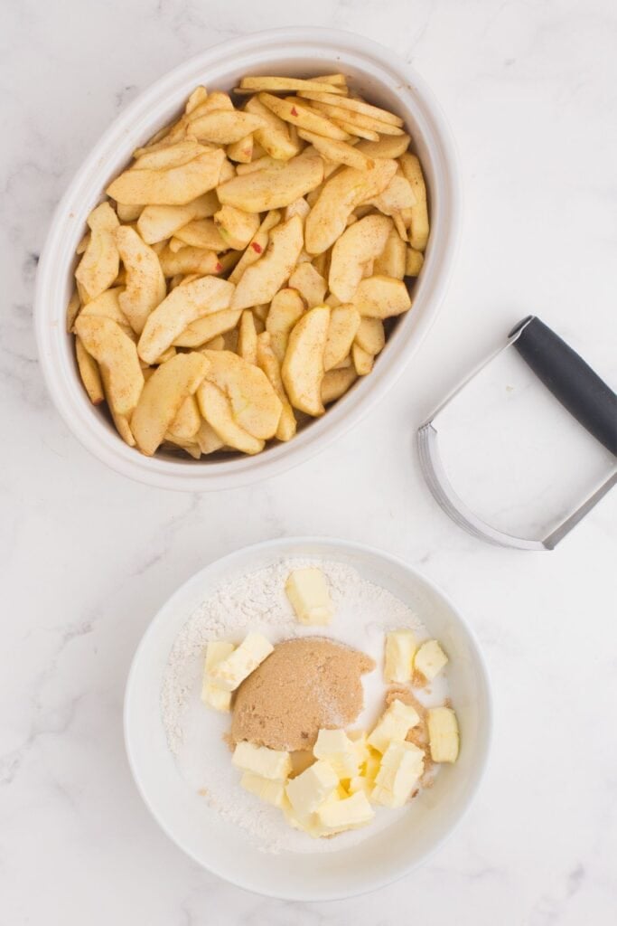 Sliced apples in a large serving dish next to a bowl with sugar, flour, brown sugar, and butter. A pastry cutter is set in between the two bowls.