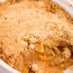 Completed apple crisp without oats in a white serving dish. A serving has been removed to show the baked apples inside.