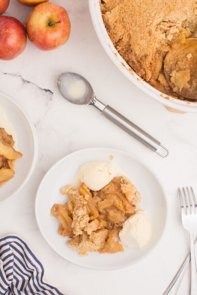 A serving of apple crisp in a white bowl with two scoops of ice cream. A ice cream scoops sits right next to the plate along with the serving dish of the baked apple crisp.