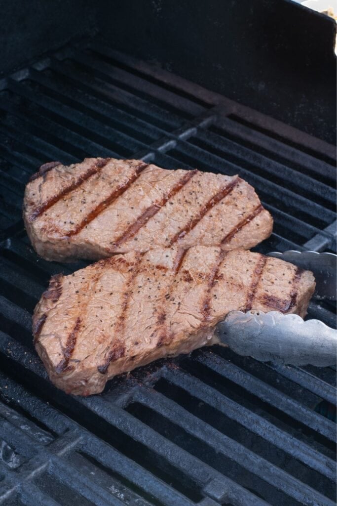 Two steaks on the grill. Tongs are flipping a steak.