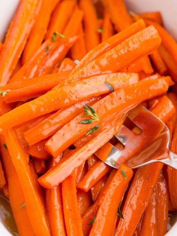 Maple glazed carrots in a white serving dish. A serving fork is holding a serving of carrots.