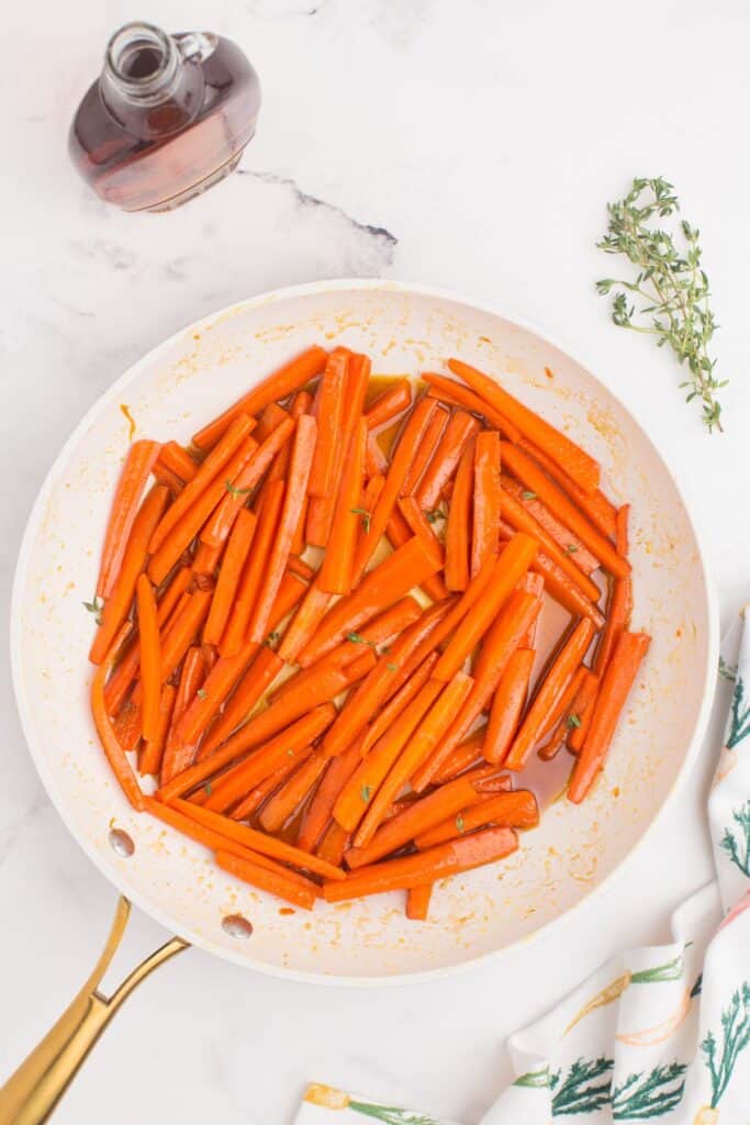 A skillet with glazed carrots cooking in it. A sprig of thyme and a bottle of maple syrup are placed next to the skillet.