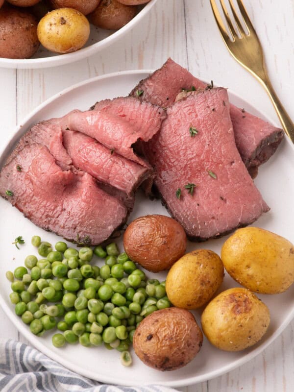 Thinly shaved slices of bottom round roast on a white dinner plate with peas and baby potatoes.