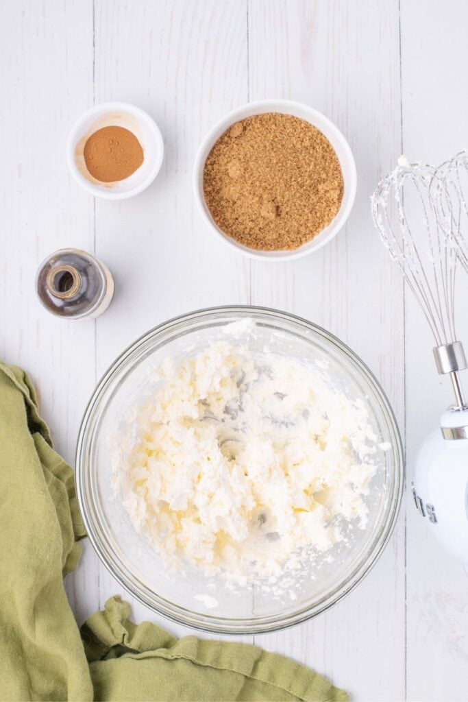 A glass bowl with whipped cream cheese in it. A hand mixer is placed just to the right of the bowl. There are two small bowls of brown sugar and cinnamon above the glass bowl with a bottle of vanilla.