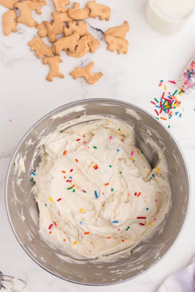 Mixing bowl with Funfetti Dip. Bowl is surrounded by animal crackers, a glass of milk, and rainbow sprinkles.