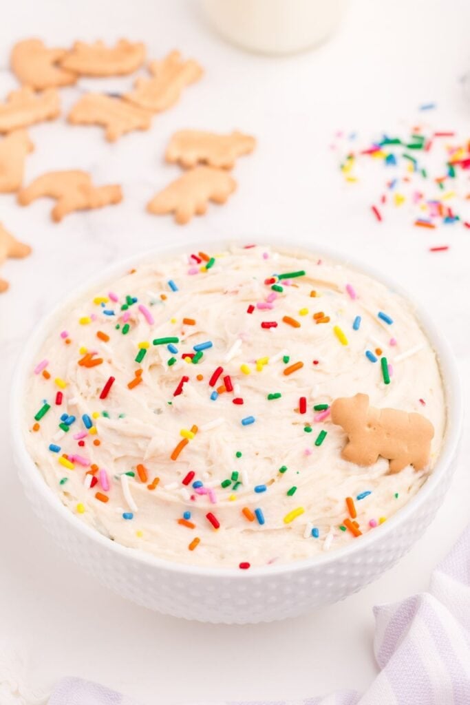 Completed Funfetti dip in a white bowl. Dip is decorated with rainbow sprinkles and a single animal cracker.