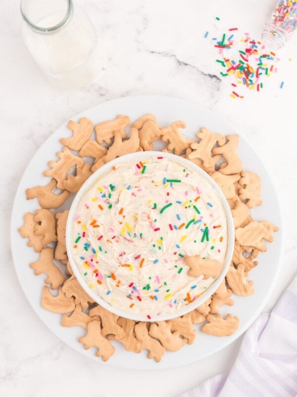Completed Funfetti dip in a white bowl on a white plate with animal crackers surrounding the bowl.