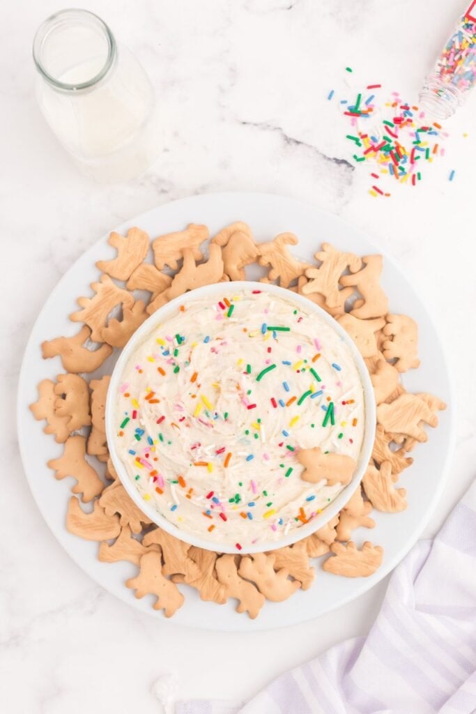 Completed Funfetti dip in a white bowl on a white plate with animal crackers surrounding the bowl.