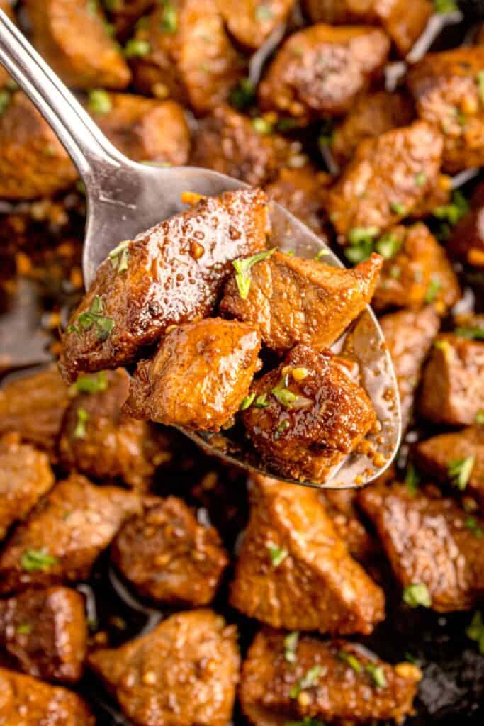 A serving spoon is lifting a portion of steak bites above the cast iron pan.