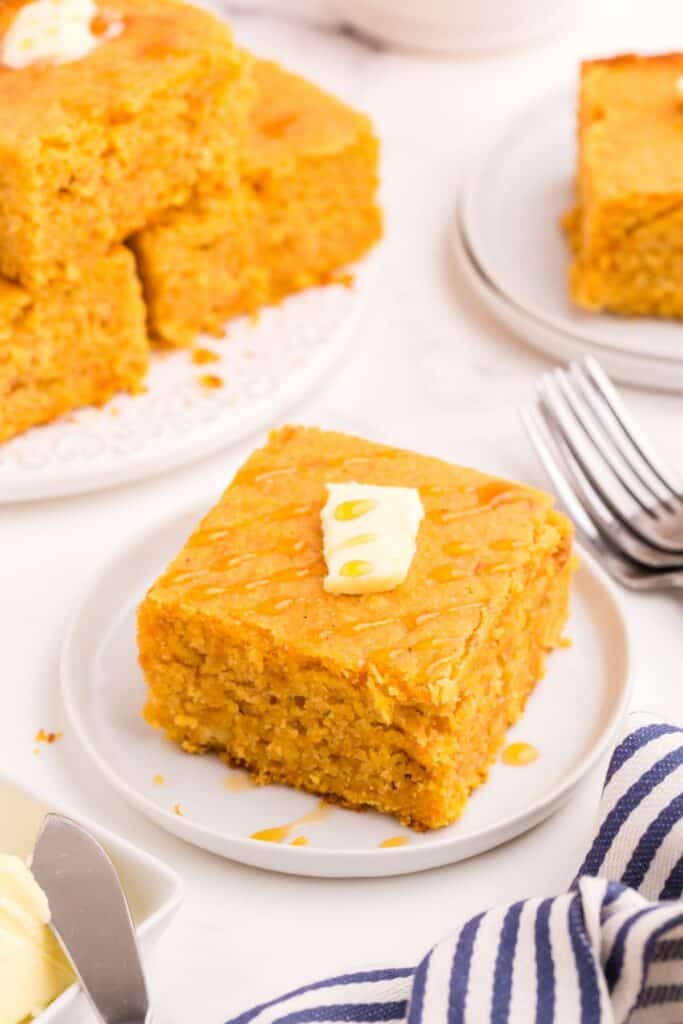 A slice of sweet potato cornbread on a small white plate. Cornbread has a pat of butter on top and a drizzle of honey.