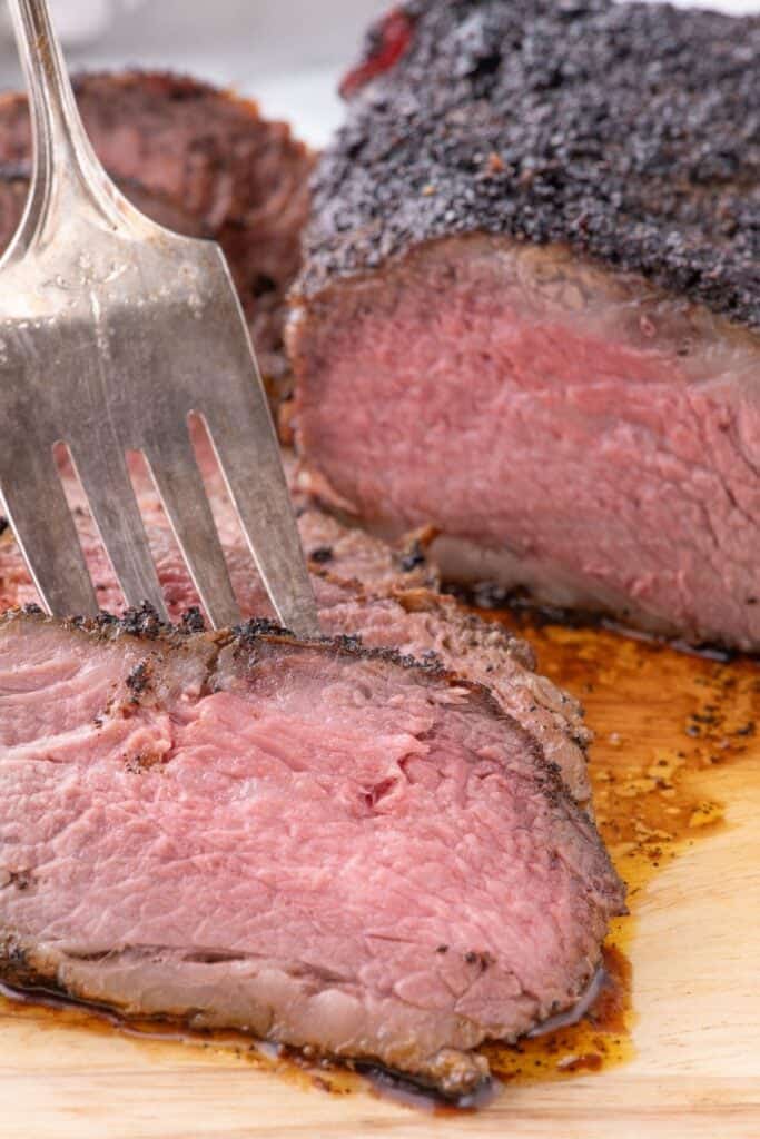 Large serving fork lifting a slice of tri tip off a stack of slices. The rest of the unsliced tri tip is right next to the slices on a wooden cutting board.