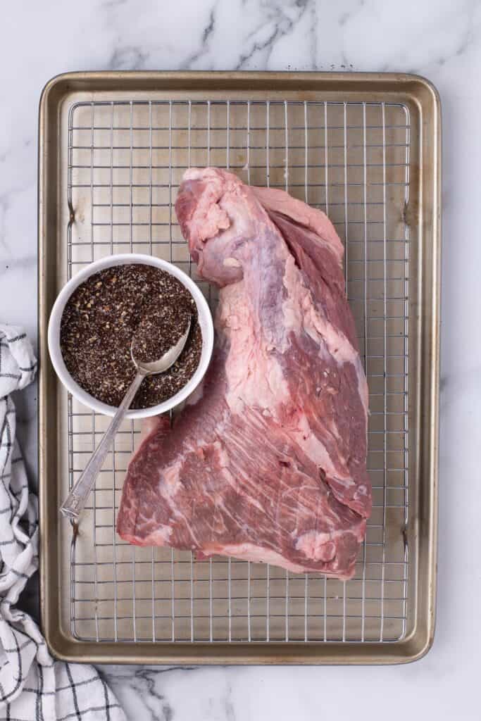 Raw tri tip cut on a baking sheet with a wire rack. A bowl of homemade seasoning is set next to the tri tip.