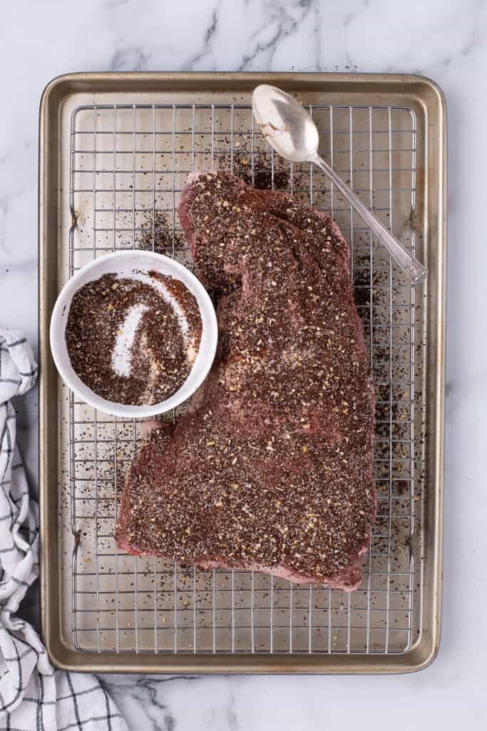 Raw tri tip cut heavily seasoned with homemade seasoning over a baking sheet with a wire rack.