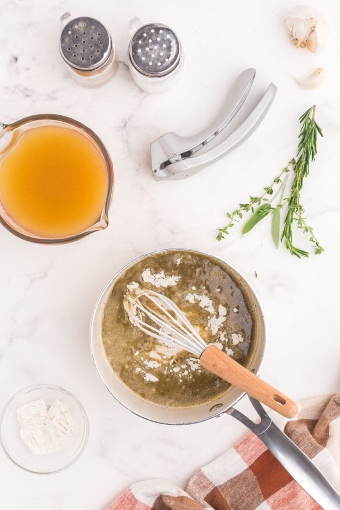 Saucepan with butter, flour, and spices being whisked into a slurry. Saucepan is surrounded by fresh herbs, garlic, a garlic crusher, salt and pepper shakers, a measuring cup of turkey broth, and a small bowl of flour.