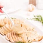 White serving dish with three sliced turkey breasts. A spoon is pouring gravy over the top of the turkey breasts.