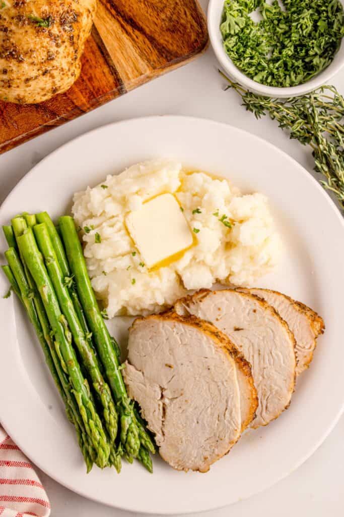A white dinner plate with sliced turkey breast, roasted asparagus, and mashed potatoes with butter.