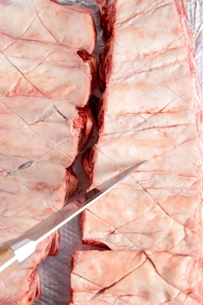 A knife is scoring the fat side of the uncooked beef ribs.