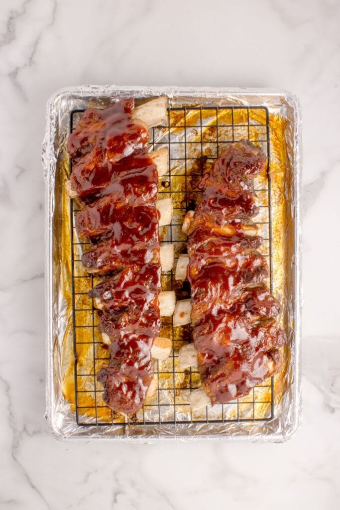 Two cooked racks of beef ribs resting on a foil-lined baking sheet with a wire rack.