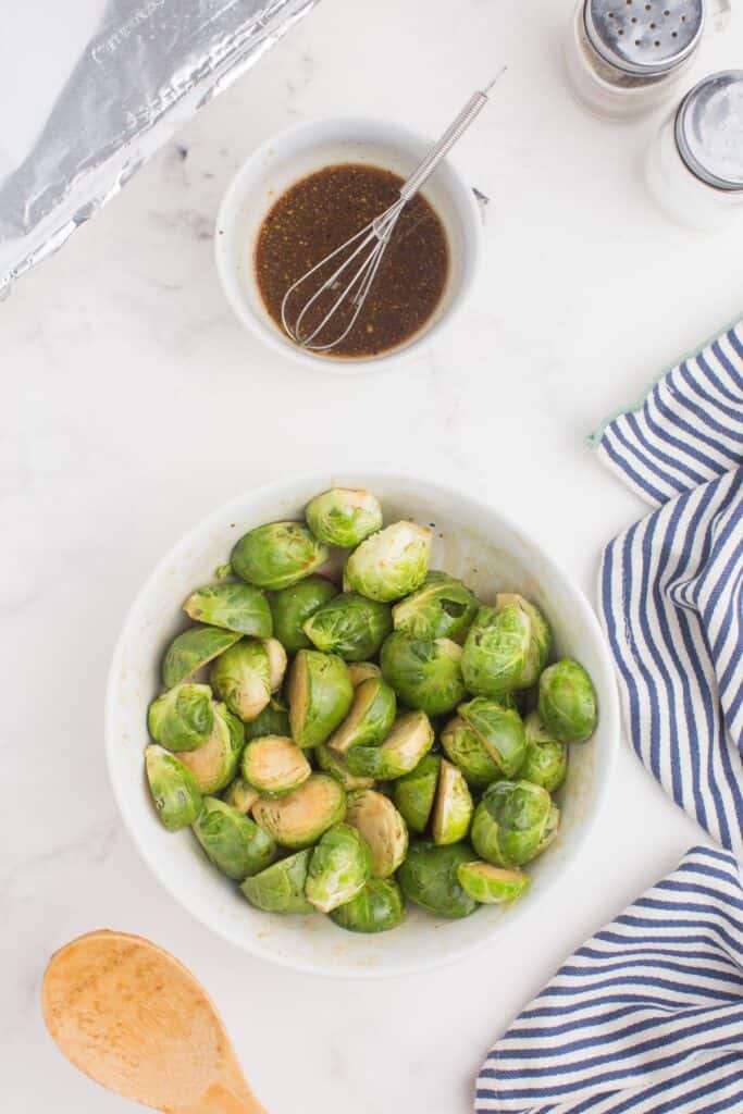 Brussels sprouts in a white bowl, tossed with half of the glaze recipe. The remaining glaze is in a white bowl placed next to the brussels sprouts.