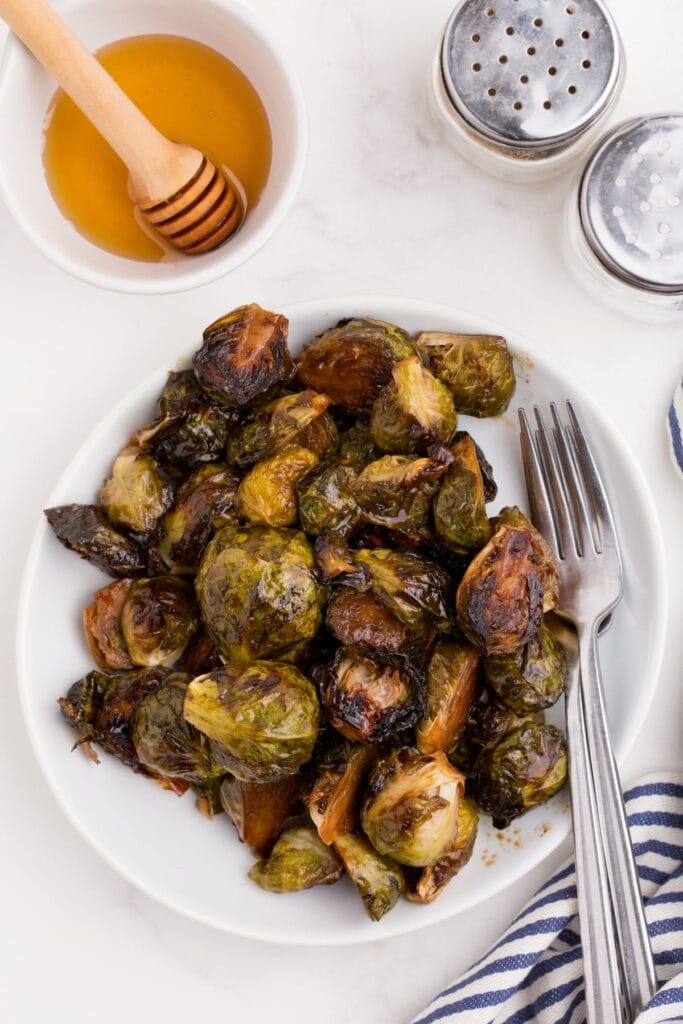 Finished brussels sprouts recipe on a white dinner plate. A small bowl of honey is beside the plate along with a pair of salt and pepper shakers.