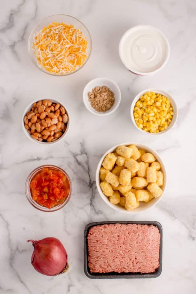 Ingredients for Taco Tater Tot Casserole: ground beef, onion, corn, pinto beans, Rotel, sour cream, Mexican cheese, taco seasoning, and tater tots.