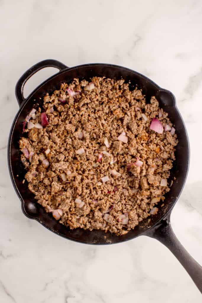 A cast iron pan with cooked and crumbled ground beef and a diced red onion.
