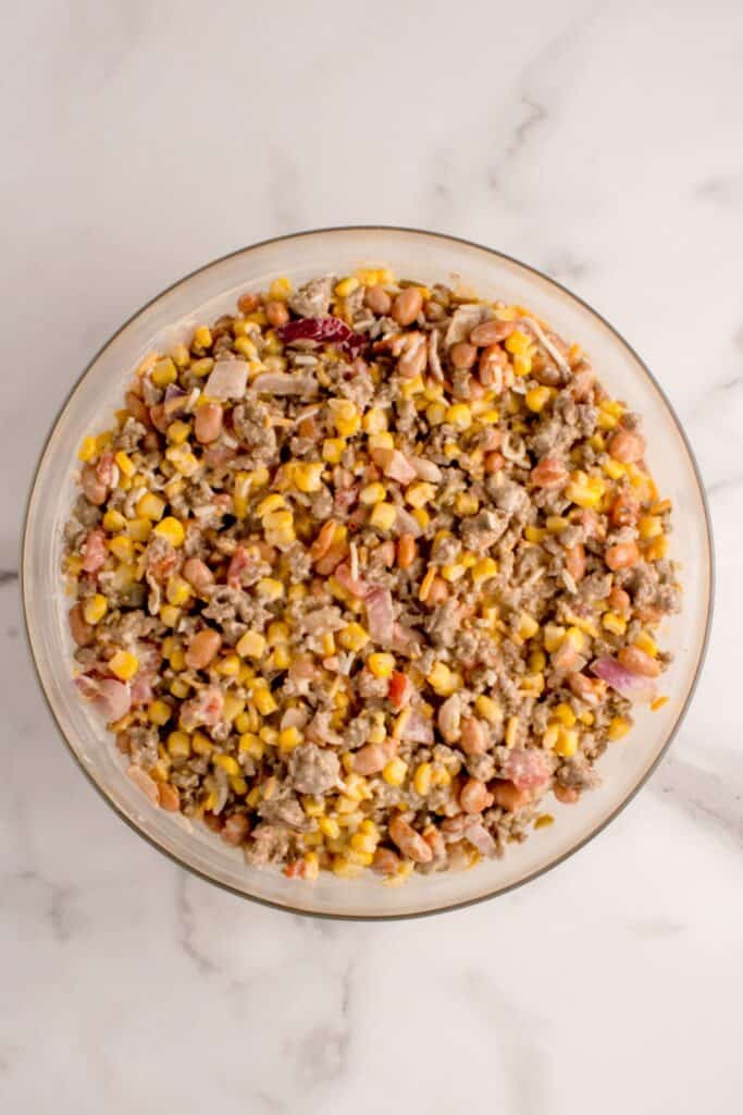 Glass mixing bowl with corn, cheese, Rotel, pinto beans, and sour cream mixed together.