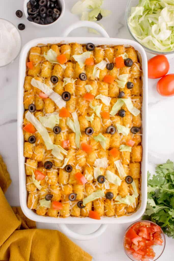 Finished taco tater tot casserole in a white baking dish with lettuce, sliced black olives, and diced tomatoes on top.