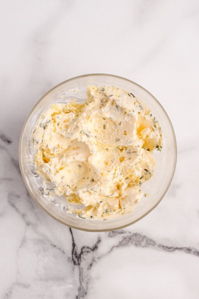 Butter mixture in a glass mixing bowl with unsalted butter, fresh rosemary, fresh thyme, garlic, salt, and ground black pepper.