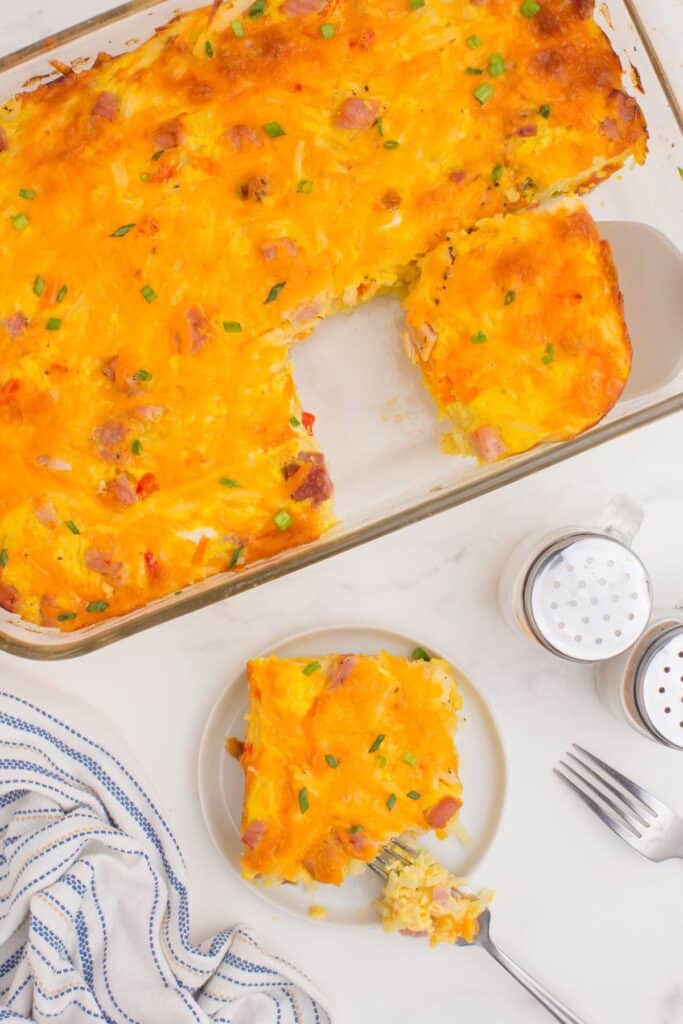 Completed ham and egg casserole in a glass baking dish. One slice has been cut out and placed on a small white plate. A fork has pulled off a bite.