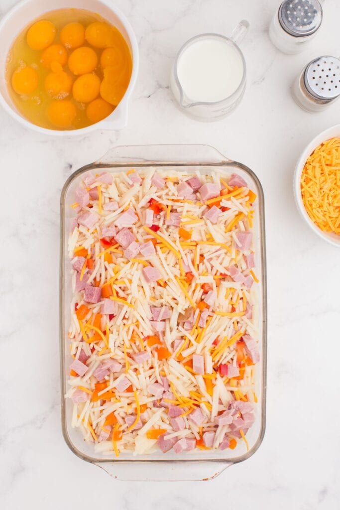 Filling ingredients prepped in a glass baking dish. A mixing bowl with eggs is above it along with a small pitcher of milk.