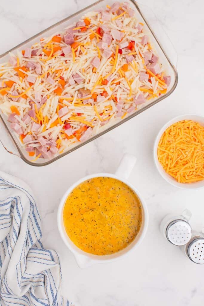 Egg mixture is combined in a separate bowl, next to a medium sized bowl of shredded cheese and a glass baking dish with hash browns, chopped ham, and bell pepper.
