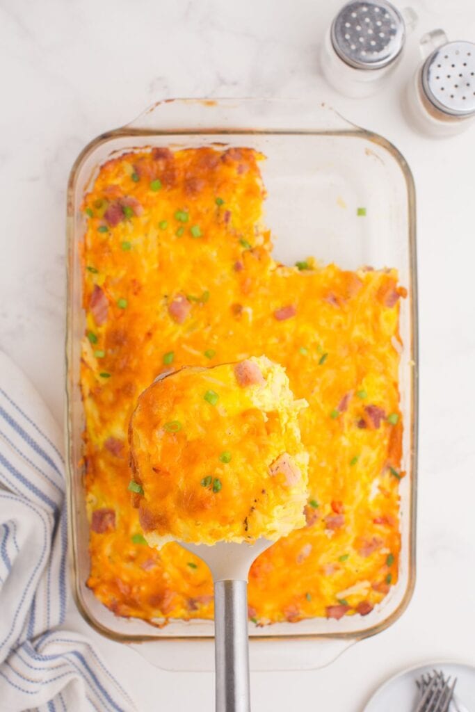 Baked ham and egg casserole in a glass baking dish. A spatula is lifting a single serving above the casserole.