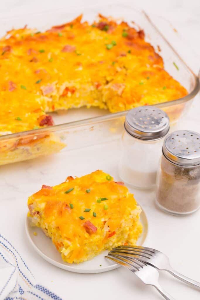 Ham and egg casserole in a glass baking dish. One slice has been cut out and placed on a small white plate.