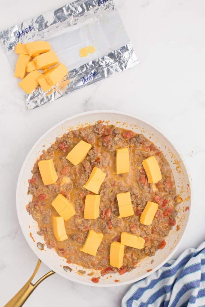 Ground beef and Rotel mixture in a large skillet with chucks of Velveeta cheese being melted into it.