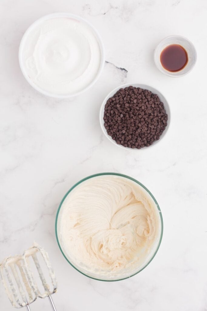 A glass mixing bowl with a whipped cream cheese mixture in it. A bowl of chocolate chips, pinch bowl of vanilla, and tub of Cool Whip sit next to the bowl.