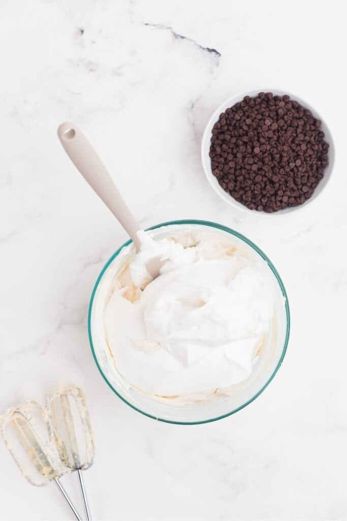 A glass mixing bowl with a whipped cream cheese mixture in it and Cool Whip on top. A bowl of chocolate chips sits next to the bowl.