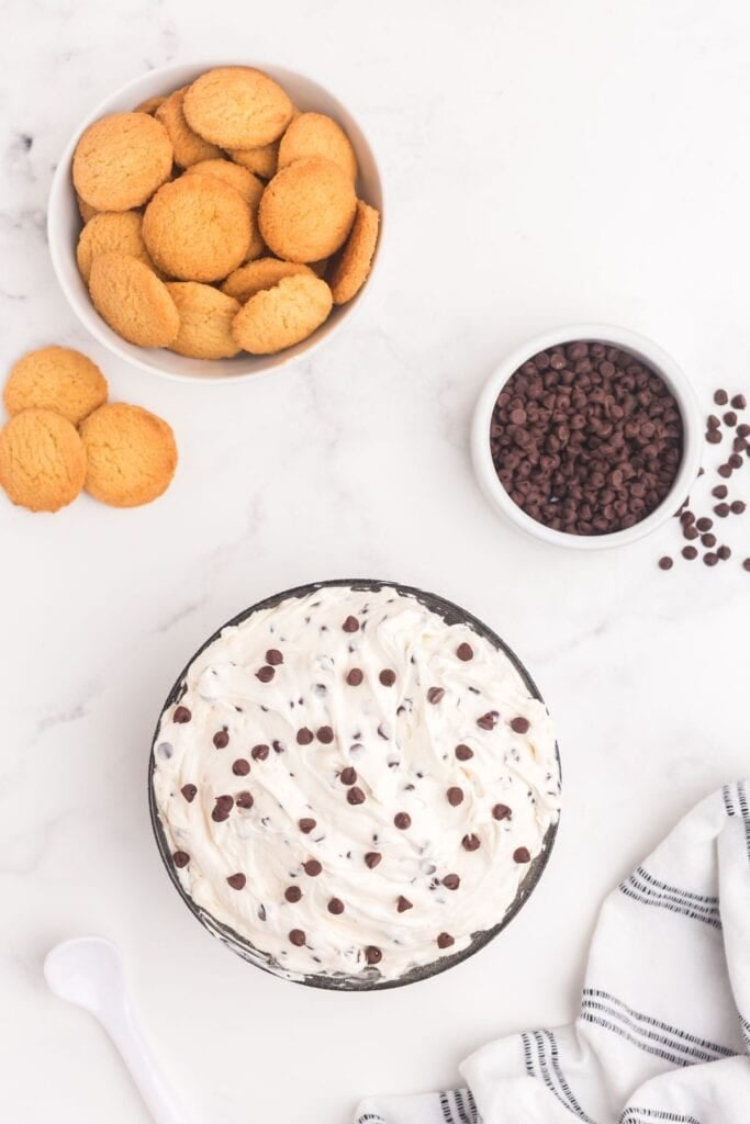 A bowl of cookie dough dip next to two other bowls, one of chocolate chips and one of vanilla cookies.