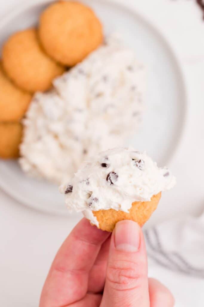 A cookie with a scoop of cookie dough dip on it, held above a white plate with a serving of dip and cookies.