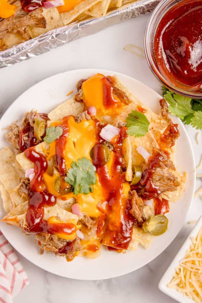 Pulled pork nachos on a white dinner plate. A bowl BBQ sauce and the platter of nachos is next to the plate.